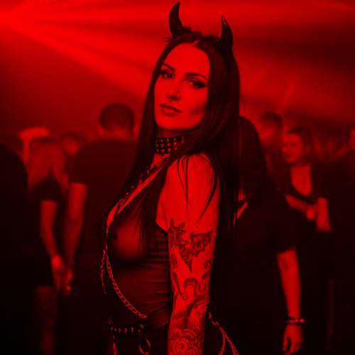 A woman with long dark hair, wearing a black devil horn headband, a spiked choker, a leather-look top with chains, and dark pants, in a dimly lit red club scene; tattoos on her arm and nose ring visible.